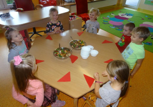 Grupa dzieci siedzi z uśmiechem przy stole nakrytym serwetkami a na środku stoją miski z pokrojonymi owocami.
